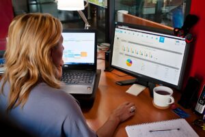A woman at a desk using a laptop and a monitor displaying Wit smart building data charts and analytics.