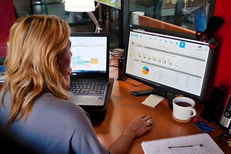 A woman at a desk using a laptop and a monitor displaying Wit smart building data charts and analytics.
