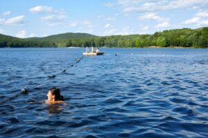 A person swims in a blue lake near a buoy line and a floating wooden dock with forested hills in the background.