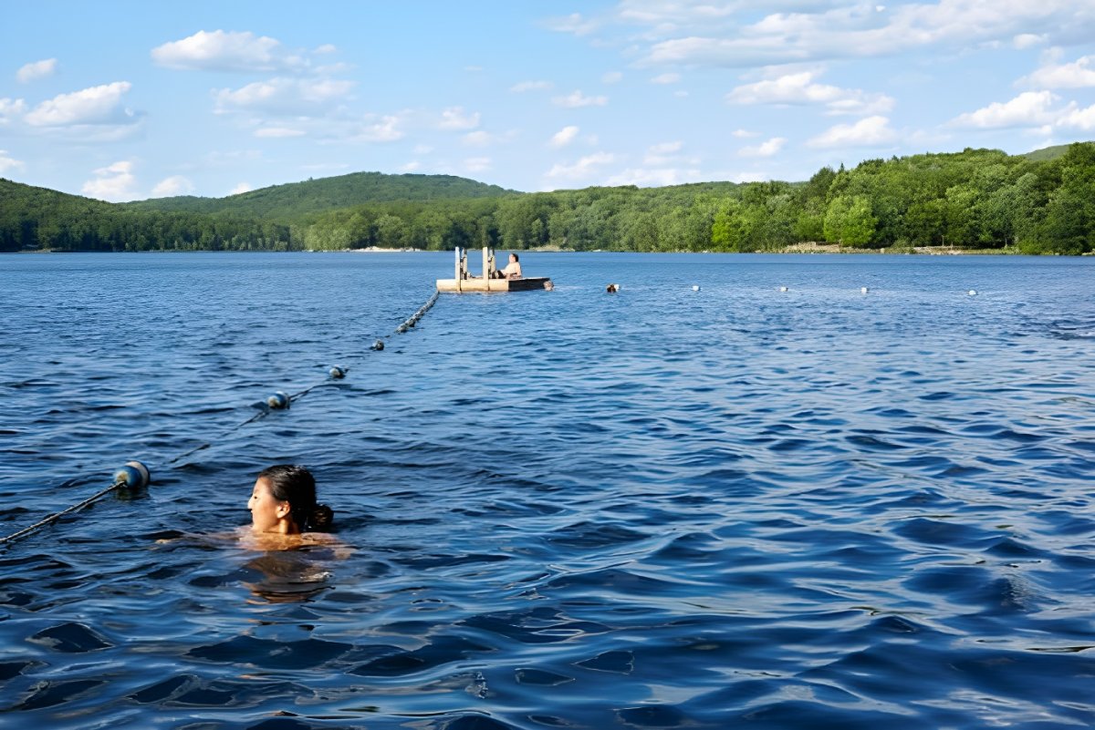 A person swims in a blue lake near a buoy line and a floating wooden dock with forested hills in the background.